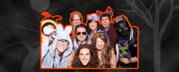 Group of people at a costume party, wearing a variety of outfits including a superhero and an animal onesie, smiling for the camera.