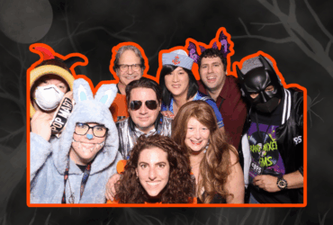 Group of people at a costume party, wearing a variety of outfits including a superhero and an animal onesie, smiling for the camera.