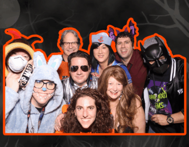 Group of people at a costume party, wearing a variety of outfits including a superhero and an animal onesie, smiling for the camera.