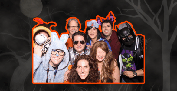 Group of people at a costume party, wearing a variety of outfits including a superhero and an animal onesie, smiling for the camera.