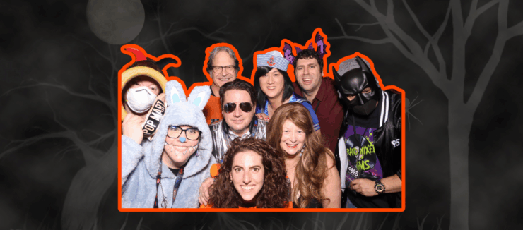 Group of people at a costume party, wearing a variety of outfits including a superhero and an animal onesie, smiling for the camera.