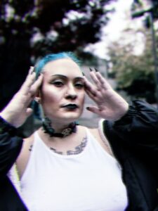Person with blue hair and a visible tattoo on their neck standing outdoors, holding their hands near their face, expression focused.