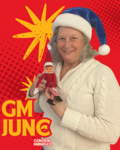 A person wearing a Santa hat holding an Elf on the Shelf toy, with text labels "GM June" and "General Manager" on a red, dotted background.