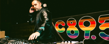 DJ performing at a mixing console with a colorful "c895" logo in the background.