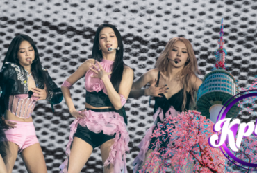 Four performers are on stage, dancing energetically against a black and white patterned backdrop. They wear stylish pink and black outfits. A graphic in the foreground features cherry blossoms and a tower, with the text "Kplus" inside a circle.