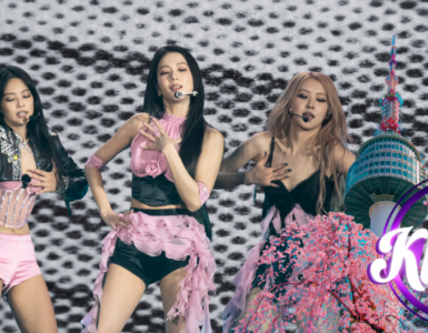 Four performers are on stage, dancing energetically against a black and white patterned backdrop. They wear stylish pink and black outfits. A graphic in the foreground features cherry blossoms and a tower, with the text "Kplus" inside a circle.