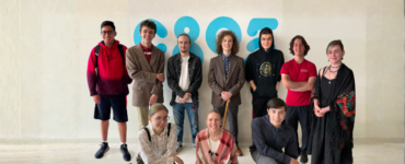 Six people stand and pose together in a room with various posters and schedules on the wall. Some of them are smiling and giving thumbs up.