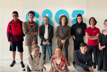 Six people stand and pose together in a room with various posters and schedules on the wall. Some of them are smiling and giving thumbs up.