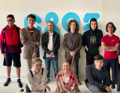 Six people stand and pose together in a room with various posters and schedules on the wall. Some of them are smiling and giving thumbs up.