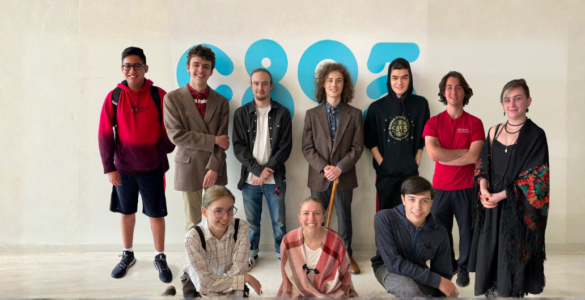 Six people stand and pose together in a room with various posters and schedules on the wall. Some of them are smiling and giving thumbs up.
