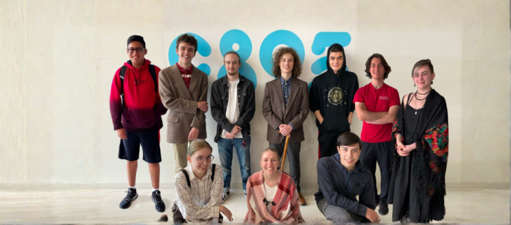 Six people stand and pose together in a room with various posters and schedules on the wall. Some of them are smiling and giving thumbs up.