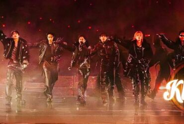 A group of performers dressed in dark clothing and sunglasses are energetically posing on stage. The image features dramatic lighting and a smoky atmosphere. The word "Kplus" is displayed in the lower right corner.