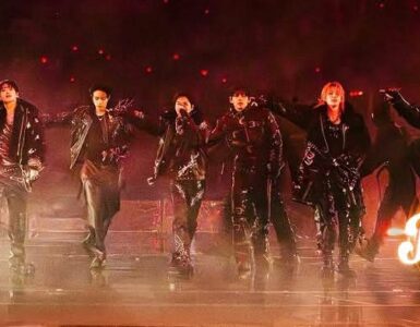 A group of performers dressed in dark clothing and sunglasses are energetically posing on stage. The image features dramatic lighting and a smoky atmosphere. The word "Kplus" is displayed in the lower right corner.