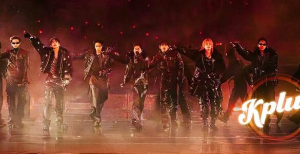 A group of performers dressed in dark clothing and sunglasses are energetically posing on stage. The image features dramatic lighting and a smoky atmosphere. The word "Kplus" is displayed in the lower right corner.