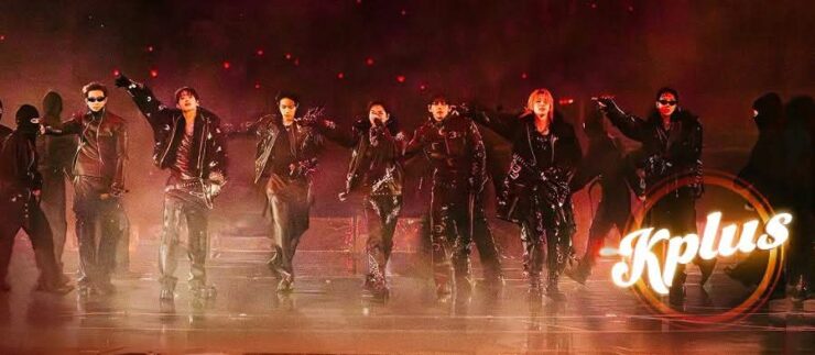 A group of performers dressed in dark clothing and sunglasses are energetically posing on stage. The image features dramatic lighting and a smoky atmosphere. The word "Kplus" is displayed in the lower right corner.