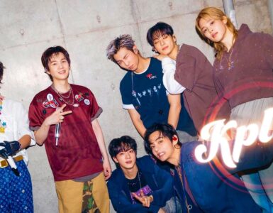 A group of seven people posing energetically in front of a concrete wall. They are dressed in stylish, casual outfits and are smiling or making playful gestures. The word "Kplus" is prominently displayed in the bottom right corner.