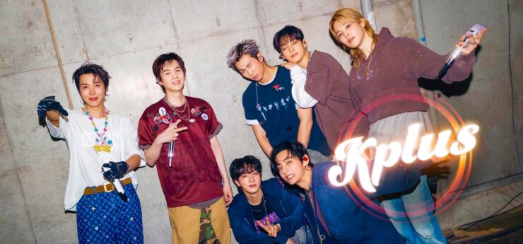 A group of seven people posing energetically in front of a concrete wall. They are dressed in stylish, casual outfits and are smiling or making playful gestures. The word "Kplus" is prominently displayed in the bottom right corner.