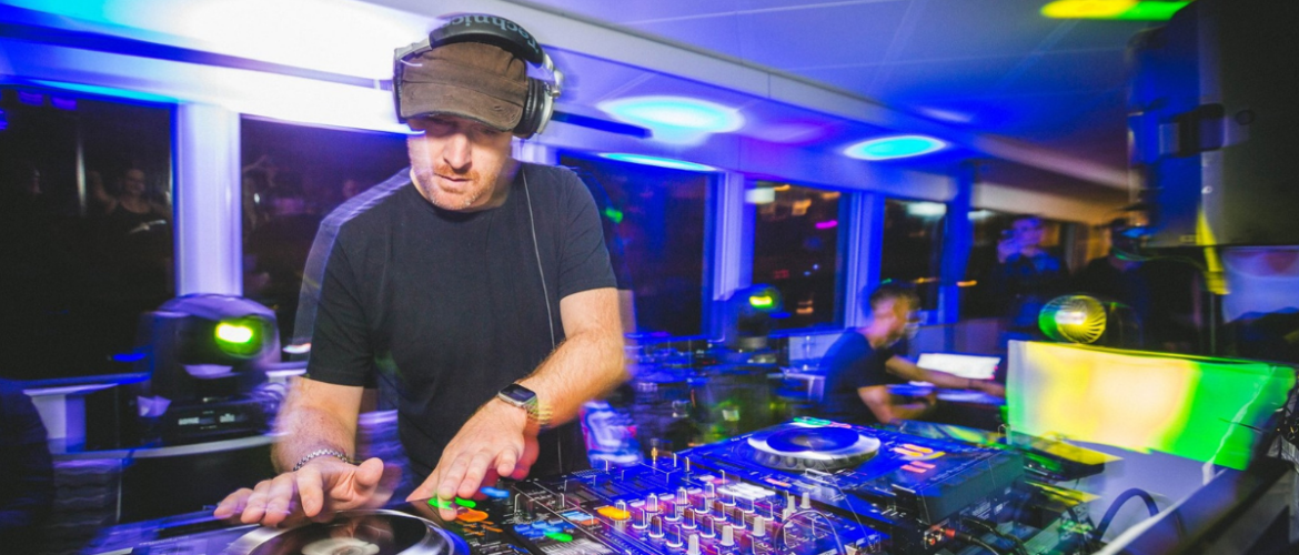 A person is DJing at a vibrant event. The setup includes turntables and mixer equipment. Colorful lights create an energetic atmosphere in the room filled with people.