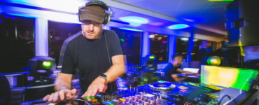 A person is DJing at a vibrant event. The setup includes turntables and mixer equipment. Colorful lights create an energetic atmosphere in the room filled with people.