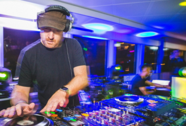 A person is DJing at a vibrant event. The setup includes turntables and mixer equipment. Colorful lights create an energetic atmosphere in the room filled with people.