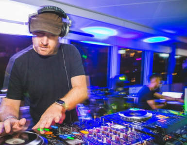 A person is DJing at a vibrant event. The setup includes turntables and mixer equipment. Colorful lights create an energetic atmosphere in the room filled with people.