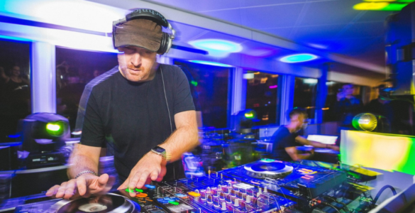 A person is DJing at a vibrant event. The setup includes turntables and mixer equipment. Colorful lights create an energetic atmosphere in the room filled with people.