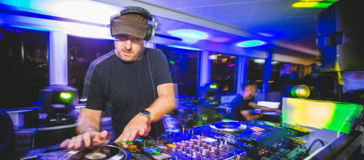 A person is DJing at a vibrant event. The setup includes turntables and mixer equipment. Colorful lights create an energetic atmosphere in the room filled with people.