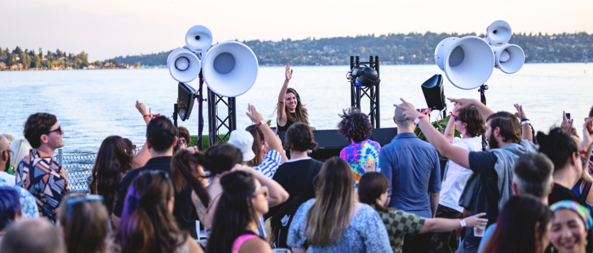 A lively crowd enjoying an outdoor music event by a lake, featuring a DJ on stage with large speakers. The background showcases a scenic view of the lake and a distant shoreline with trees.