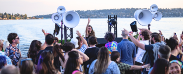 A lively crowd enjoying an outdoor music event by a lake, featuring a DJ on stage with large speakers. The background showcases a scenic view of the lake and a distant shoreline with trees.