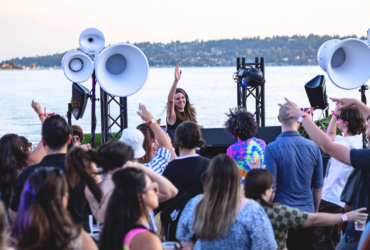 A lively crowd enjoying an outdoor music event by a lake, featuring a DJ on stage with large speakers. The background showcases a scenic view of the lake and a distant shoreline with trees.