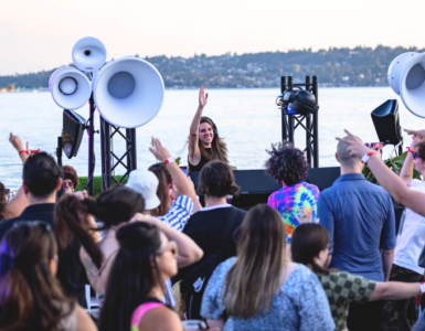 A lively crowd enjoying an outdoor music event by a lake, featuring a DJ on stage with large speakers. The background showcases a scenic view of the lake and a distant shoreline with trees.