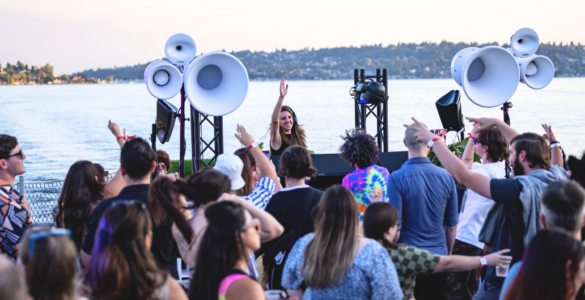 A lively crowd enjoying an outdoor music event by a lake, featuring a DJ on stage with large speakers. The background showcases a scenic view of the lake and a distant shoreline with trees.