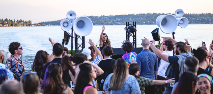 A lively crowd enjoying an outdoor music event by a lake, featuring a DJ on stage with large speakers. The background showcases a scenic view of the lake and a distant shoreline with trees.
