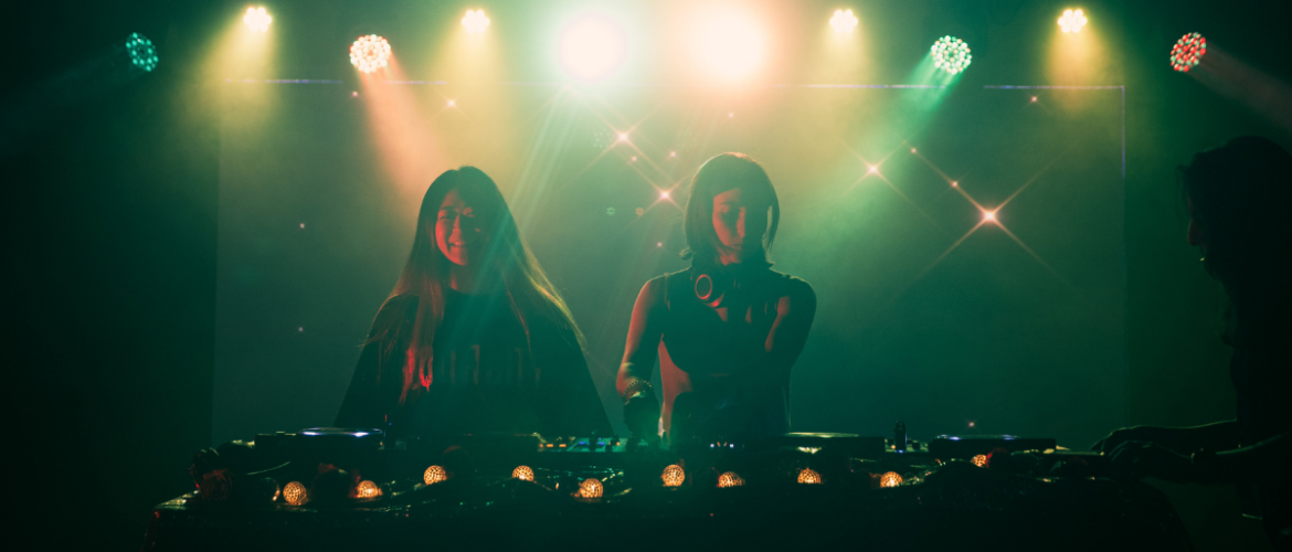Two individuals are DJing on stage, illuminated by colorful lights. A third person stands nearby, slightly obscured. The atmosphere is vibrant, with a backdrop of sparkling lights, creating a lively setting.