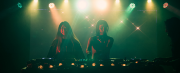 Two individuals are DJing on stage, illuminated by colorful lights. A third person stands nearby, slightly obscured. The atmosphere is vibrant, with a backdrop of sparkling lights, creating a lively setting.