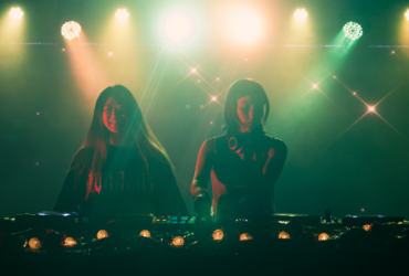 Two individuals are DJing on stage, illuminated by colorful lights. A third person stands nearby, slightly obscured. The atmosphere is vibrant, with a backdrop of sparkling lights, creating a lively setting.