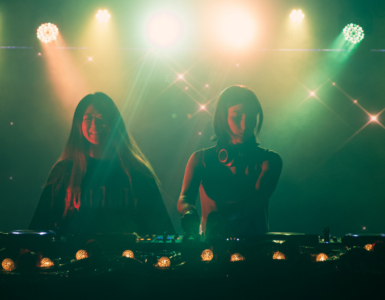 Two individuals are DJing on stage, illuminated by colorful lights. A third person stands nearby, slightly obscured. The atmosphere is vibrant, with a backdrop of sparkling lights, creating a lively setting.