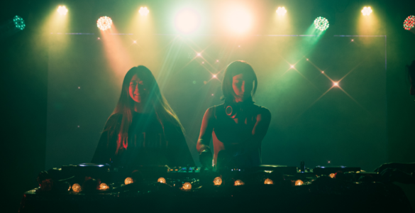 Two individuals are DJing on stage, illuminated by colorful lights. A third person stands nearby, slightly obscured. The atmosphere is vibrant, with a backdrop of sparkling lights, creating a lively setting.
