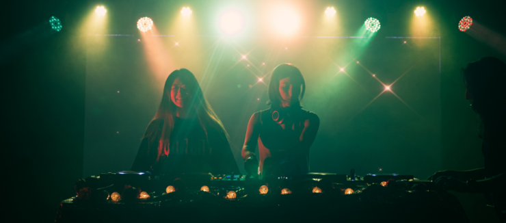 Two individuals are DJing on stage, illuminated by colorful lights. A third person stands nearby, slightly obscured. The atmosphere is vibrant, with a backdrop of sparkling lights, creating a lively setting.
