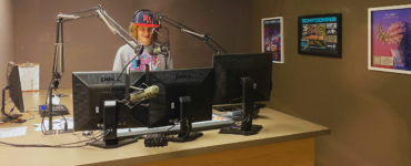 Radio host standing at a studio desk with two computer monitors, wearing a cap with the letters "NF". A microphone is positioned in front of them. Posters are visible on the wall in the background.