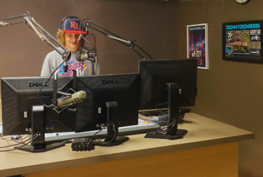 Radio host standing at a studio desk with two computer monitors, wearing a cap with the letters "NF". A microphone is positioned in front of them. Posters are visible on the wall in the background.