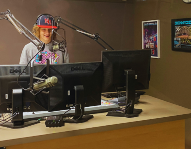 Radio host standing at a studio desk with two computer monitors, wearing a cap with the letters "NF". A microphone is positioned in front of them. Posters are visible on the wall in the background.