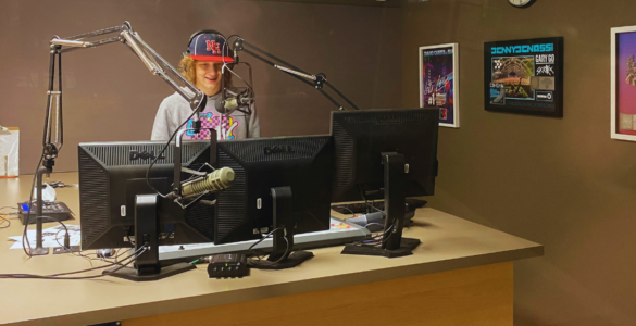 Radio host standing at a studio desk with two computer monitors, wearing a cap with the letters "NF". A microphone is positioned in front of them. Posters are visible on the wall in the background.