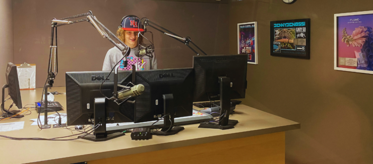 Radio host standing at a studio desk with two computer monitors, wearing a cap with the letters "NF". A microphone is positioned in front of them. Posters are visible on the wall in the background.