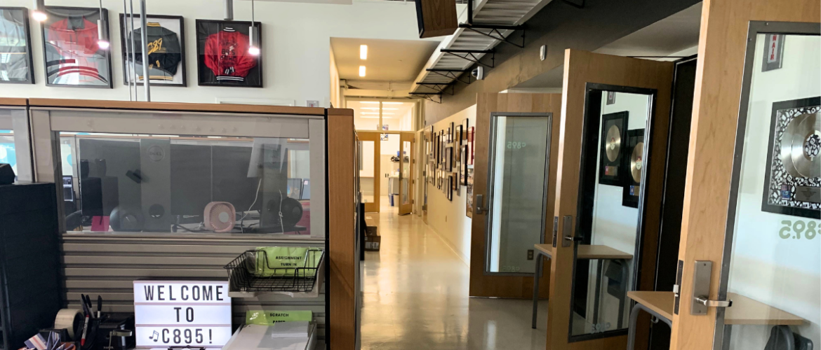 A modern office space featuring a hallway lined with various framed pictures. To the left, a glass partition displays a sign reading "Welcome to C89.5!" Above, sports jerseys are framed and hung on the wall. The space is well-lit and organized.