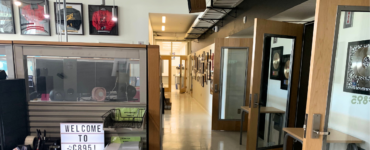 A modern office space featuring a hallway lined with various framed pictures. To the left, a glass partition displays a sign reading "Welcome to C89.5!" Above, sports jerseys are framed and hung on the wall. The space is well-lit and organized.
