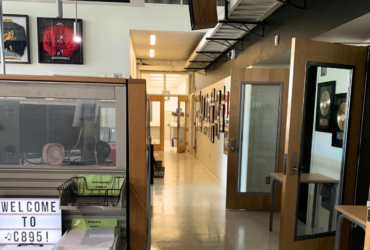 A modern office space featuring a hallway lined with various framed pictures. To the left, a glass partition displays a sign reading "Welcome to C89.5!" Above, sports jerseys are framed and hung on the wall. The space is well-lit and organized.