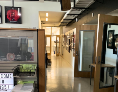 A modern office space featuring a hallway lined with various framed pictures. To the left, a glass partition displays a sign reading "Welcome to C89.5!" Above, sports jerseys are framed and hung on the wall. The space is well-lit and organized.
