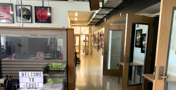 A modern office space featuring a hallway lined with various framed pictures. To the left, a glass partition displays a sign reading "Welcome to C89.5!" Above, sports jerseys are framed and hung on the wall. The space is well-lit and organized.