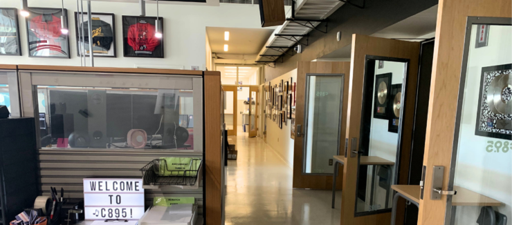 A modern office space featuring a hallway lined with various framed pictures. To the left, a glass partition displays a sign reading "Welcome to C89.5!" Above, sports jerseys are framed and hung on the wall. The space is well-lit and organized.